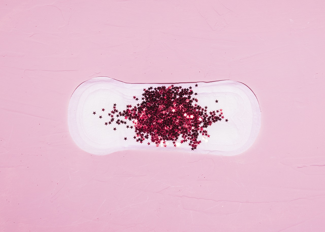 A sanitary napkin laid out on a pink background with red glitter on top to symbolize vaginal blood discharge